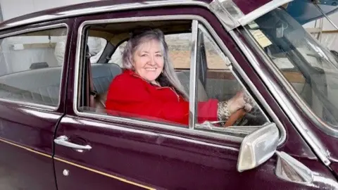 A woman in a red jacket sitting at the steering wheel of a classic car