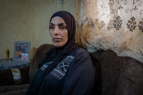 A close up of Jad's mother looking forlorn, sitting in the family living room in al-Far'a refugee camp, in the occupied West Bank. Behind her on the table is a framed picture of Jad.