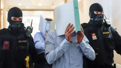 Reuters Two armed Austrian security personnel in masks flank a suspect, identified only as Beran A. The suspect is seen wearing a blue shirt and carrying a ring binder in front of his face. Photo: 28 May 2026