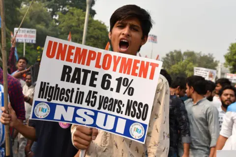 Hindustan Times via Getty Images A young man wearing a white shirt holds a placard with slogans against unemployment and looks into the camera while shouting. Behind him are other protesters