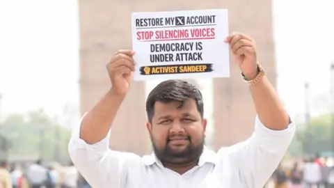 Sandeep Singh Sandeep Singh, an X user, holds up a placard saying 'Restore my X account. Stop silencing voices. Democracy under attack' on a street in India