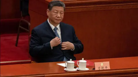 Getty Images Chinese President Xi Jinping adjusts his navy blue jacket as he sits at the closing session of the Chinese People's Political Consultative Conference (CPPCC) at the Great Hall of the People on March 11, 2026 in Beijing, China. He is sitting in front of a long wooded table with two white tea cups in front of him