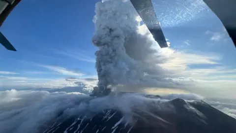 Institute of Volcanology and Seismology A large ash plume rises above a volcano. The photo appears to have been taken from a helicopter as rotor blades are in the foreground.