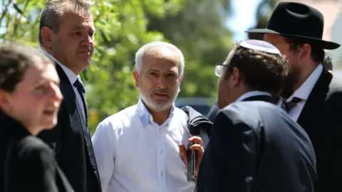Getty Images Jamal Rifi in a white button down shirt, jacket slung over his shoulder, speaks to a group of men in black suits