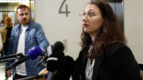 AFP via Getty Images A woman called Elisabeth Guthmann wearing a dark coat and glasses talks into microphones