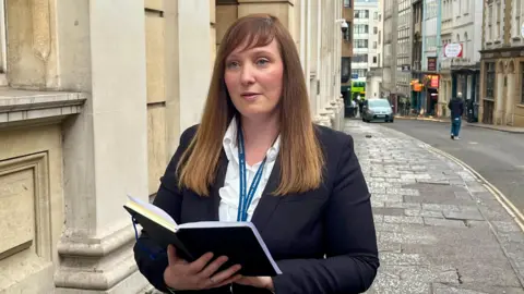 Elle MacLeod has mid-length brown hair and is wearing a white shirt and dark jacket. She is holding a notebook in her right hand which she is reading from. She is pictured on the pavement outside Bristol Crown Court