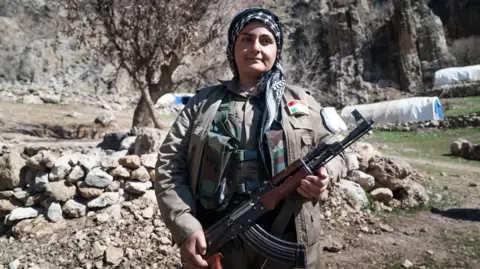 Matthew Goddard/BBC Female Iranian Kurdish fighter dressed in headscarf and grey uniform and holding an automatic weapon stands in front of a pile of stones and some tents in the distance.