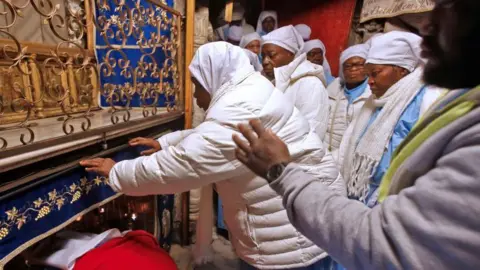 AFP via Getty Images A group of pilgrims wearing light‑coloured clothing gather inside the Grotto at the Church of the Nativity in Bethlehem. Some individuals are reaching forward to touch the ornate, blue‑draped area surrounding the entrance to the small alcove traditionally believed to mark the birthplace of Jesus Christ.