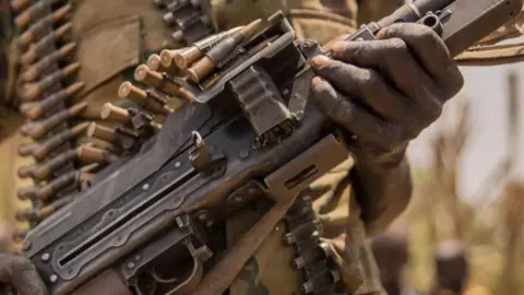 AFP via Getty Images A close-up of the body of a gun with a magazine of bullets on top of it. A hand is holding one part of the gun and more bullets can be seen, out of focus, in the background.