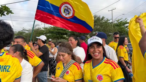 Crowd of people wearing Colombia's yellow football jersey gather on the streets, with one waving a Colombian flag and a woman smiling at the camera