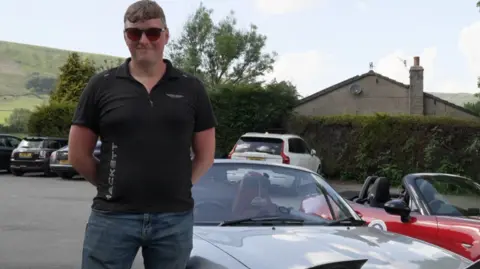 Steve Fowler wearing blue jeans, a black t shirt and sunglasses standing next to a grey convertible