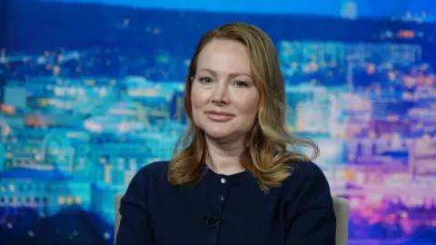 Thierry Humeau Chauntae Davies, with blonde shoulder-length hair, wearing a navy jumper, looks straight ahead. A blue and purple cityscape sits in the background.