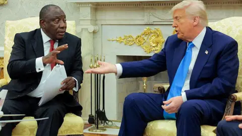 Getty Images Cyril Ramaphosa and Donald Trump sit in arm chairs in the Oval Office. Gold decorations can be seen behind them. Ramaphosa is holding a sheaf of papers in his left hand and is gesticulating to Trump who is holding out his hand.