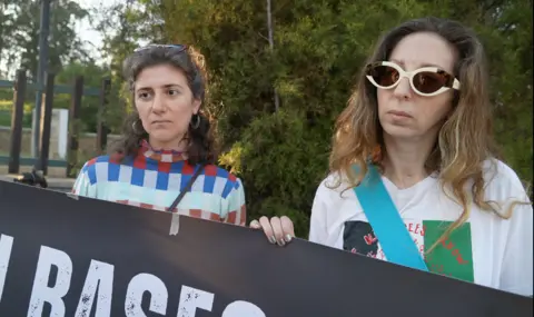 Jessica Parker/BBC Two dar-haired women hold up a black banner while looking at a person out of frame. 