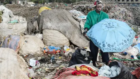 AFP via Getty Images A man in a green jacket holds a blue umbrella at a landfill site full of bails and piles of second-hand garment in Nairobi - October 2025.