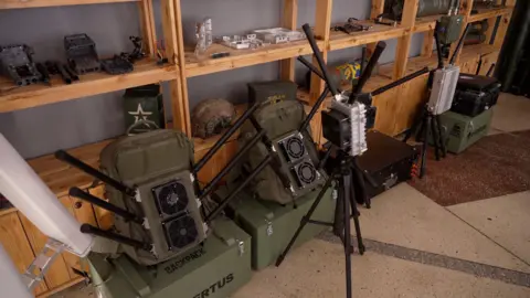 Kvertus Green anti-drone military equipment leans against a brown wooden shelving unit