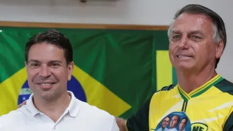 REUTERS/Ricardo Moraes Alexandre Ramagem, wearing a white Polo shirt and standing in front of a Brazilian flag, smiles at the camera. Next to him, with his hand placed on Ramagem's shoulder, is Jair Bolsonaro. He is wearing a yellow-and-green football shirt