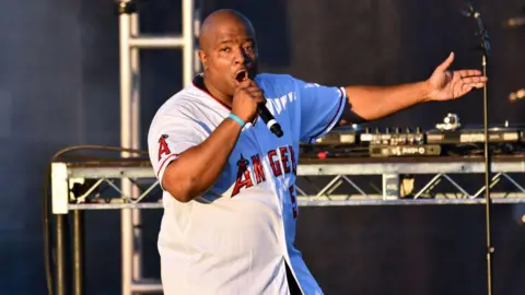 Getty Images Young MC performs onstage. He is wearing a white Los Angeles jersey with red writing and is singing into a hand-held microphone