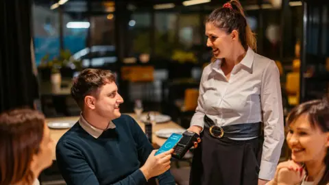 Getty Images A customer paying a bill in a restaurant, using his phone to tap against the payment device being held by a waitress