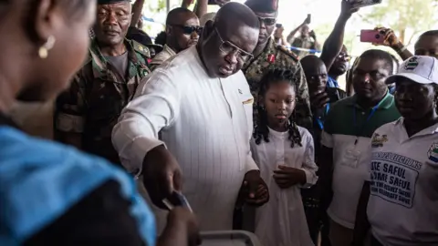 JOHN WESSELS/AFP via Getty Images A man in a white tunic with thin, pale grey, vertical stripes puts a voting paper into a box. A crowd of people stands behind him, and two men in army uniform stand close by. A girl stands next to him watching what he is doing. 