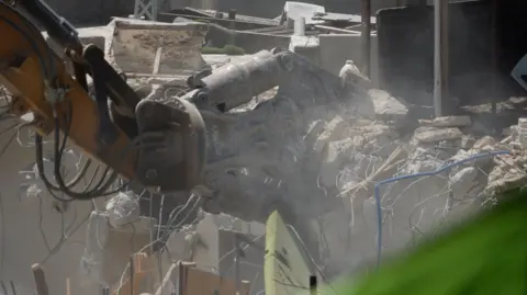 BBC An excavator picks up rubble and wires after the demolition of a building.