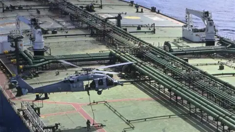 US Department of Defense A grey military helicopter hovers on top of a oil tanker, a soldier sliding down a rope towards the top deck as another uniformed soldier waits. The deck is lined with oil transportation equipment, including green pipes
