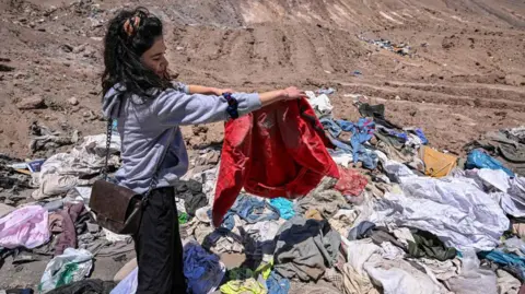 AFP via Getty Images A woman holding up clothing, a red coat, that has been dumped in a desert in northern Chile