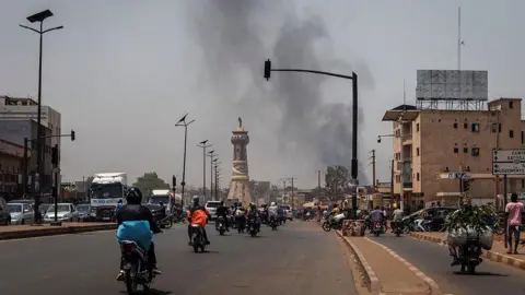 AFP via Getty Images A column of smoke rises above buildings in the distance at the end of the road. The road has dozens of motorbikes travelling along it