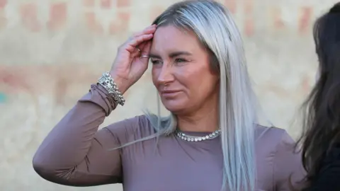 Reuters Lyanne Kennedy speaking to reporters outside Tbilisi City Court. She has long, dyed platinum-blonde hair with dark roots showing and is wearing silver necklaces, bracelets and rings. She has on a mauve, long-sleeved t-shirt. Graffiti is on the wall behind her.