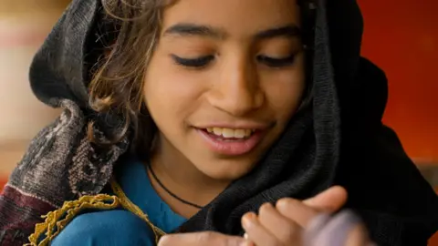 A young girl in a blue dress smiles, her eyes looking down towards her hands. She has a dark scarf with a patterned border over her head.