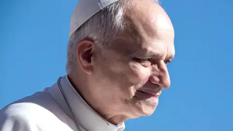 Getty Images A close-up head-and-shoulders profile image of a smiling Pope Leo XIV. A clear blue sky provides the background.