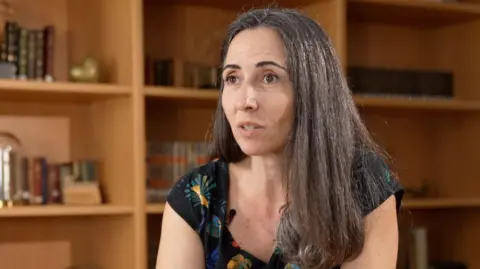 Carmona is wearing a patterned dress and has long brown hair, she is sitting near a bookshelf filled with books.