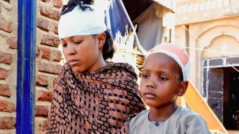 Reuters A woman and a boy injured during a drone strike in el-Obeid in North Kordofan sit outside a damaged property. Both of the heads are bandaged. They look solemn and stare away from the camera - 12 January 2026.
