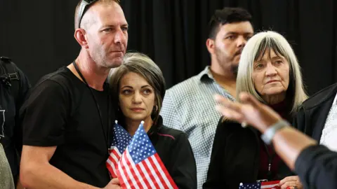 Reuters A man wearing sunglasses on his head with his arm around a woman. They are standing on the left and each holding a mini American flag. To their right, is an older woman. Behind her is a man. The background appears to be a black curtain.