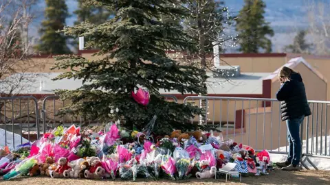 AFP via Getty Images A woman stands beside a memorial for the victims of the Tumbler Ridge shooting, set up around a spruce tree that is surrounded with flowers, teddy bears and notes. The woman has her head in her hands.