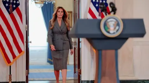Reuters Melania Trump wears a business suit with a skirt and walks toward a podium with the presidential seal at the White House