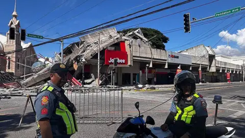 AFP via Getty Images Policemen in uniform stand in front of a barricaded road. Behind them is a partially collapsed building with a red Jollibee sign.