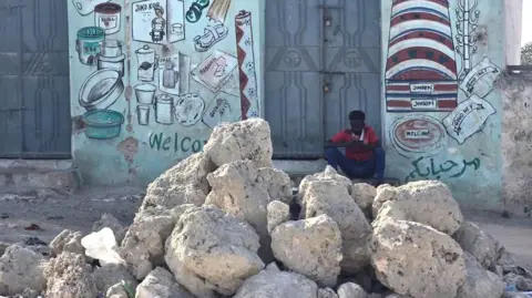 Mohamed Gabobe A pile of large stones sits in front of a locked-up shop. A man in a red T-shirt sits on the doorstep of the shop checking his phone.