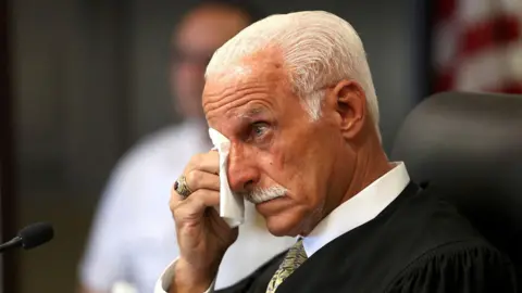 James Carbone/Pool via REUTERS A man in a judge's robe is seen in profile. He has a tan and white hair, and is wiping his eye with a tissue.