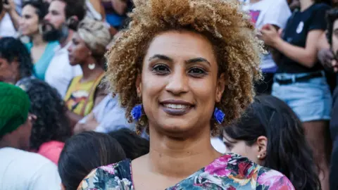 AP Marielle Franco smiling. Her hair is short, blond and curly. She is wearing a floral blouse and a pair of bright blue earings. 