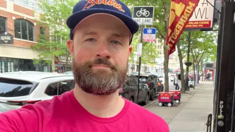 John Roberts John Roberts, wearing a pink top on a Chicago street, with two delivery robots in the background