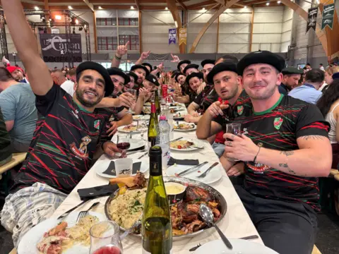 BBC A group of young men wearing matching sports tops and black berets sit at a long table with platters of food and wine
