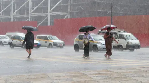 EPA/Shutterstock Three people walking across a road in heavy rain in Myanmar, with traffic in the background. All are holding umbrellas, as the rain bounces off the road crossing