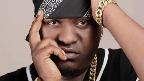 Jambomail Close-up of musician Sefu Shabani in a black bandana with a white design posing with his hands near his face. He is wearing a chunky gold chain necklace and bracelet.