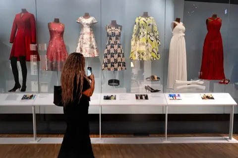 Scott Olson/Getty Images A woman views a selection of dresses worn by Michelle Obama while she was first lady. The dresses are patterned and displayed on mannequins, accompanied by the shoes and other accessories worn with them.