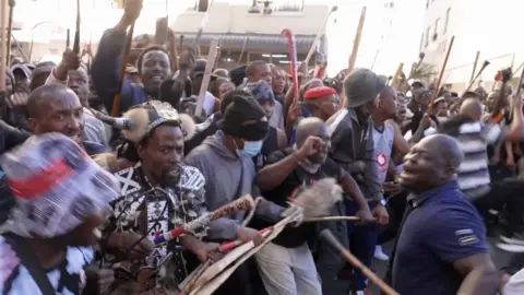 Anti-migrant protesters in South Africa - mainly men holding sticks one dressed in Zulu traditional clothing.