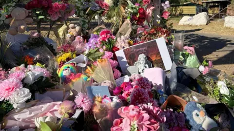 BBC: Katy Watson Bunches of flowers, soft toys and other tributes to lie in front of a wire fence 