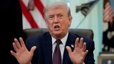 Reuters US President Donald Trump gestures with both hands while seated, with out of focus US flags in the background
