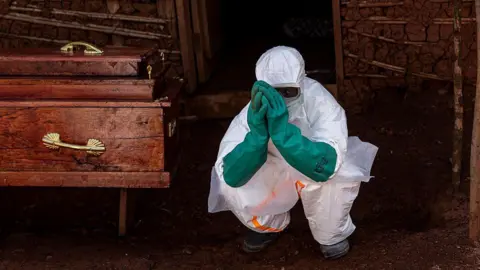 Getty Images A health worker in full PPE - white suit and green rubber gloves - crouched on the right of a wooden coffin with gold handles