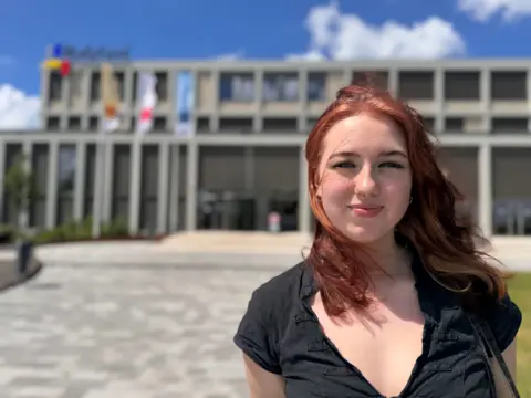 Amelie with shoulder-length red hair in the foreground, facing the camera outdoors, wearing a black short-sleeved shirt and small facial piercings. Behind, a large modern concrete building with rectangular windows and tall vertical columns spans the width of the image. Several flagpoles with light-coloured flags stand near the entrance. A wide paved area leads up to the building, bordered by low greenery. The sky is bright blue with scattered white clouds.